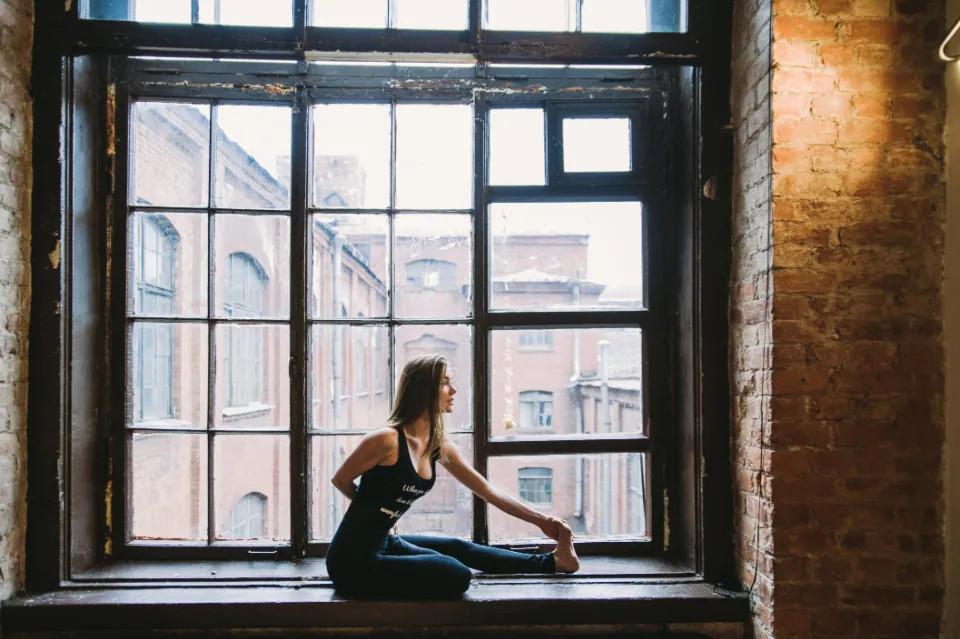 A person doing a seated hamstring and lower-back stretch by a window at home